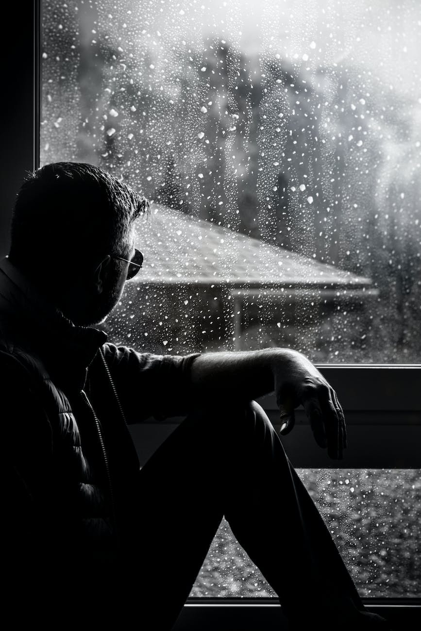 grayscale photo of man sitting beside window